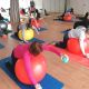 En una clase de gimnasia, los participantes se estiran mientras usan pelotas de ejercicios y pesas pequeñas. Están colocados sobre colchonetas de yoga en una sala con espejos.