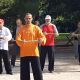Un grupo de personas practica Tai Chi al aire libre, sosteniendo palos de madera en varias posturas. El instructor, con un traje naranja, se sitúa al frente y dirige la sesión. Al fondo se ven árboles.
