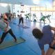 Un grupo de personas está practicando una postura de estiramiento lateral en una clase de yoga sobre colchonetas azules dentro de una sala bien iluminada con grandes ventanas y espejos.