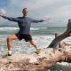 Una persona vestida con ropa deportiva se mantiene en equilibrio sobre un gran tronco de madera flotante en la playa, con los brazos extendidos, con el océano y el cielo de fondo.