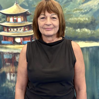 Una mujer con cabello castaño corto, presumiblemente una de las profesoras, viste una blusa negra sin mangas y está parada frente a un fondo que muestra una pagoda y un paisaje escénico.