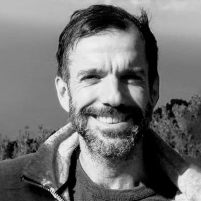 Retrato en escala de grises de un hombre con barba, sonriendo al aire libre en una zona montañosa. Reminiscencia de esos momentos atemporales compartidos por profesores mientras exploraban el terreno accidentado.